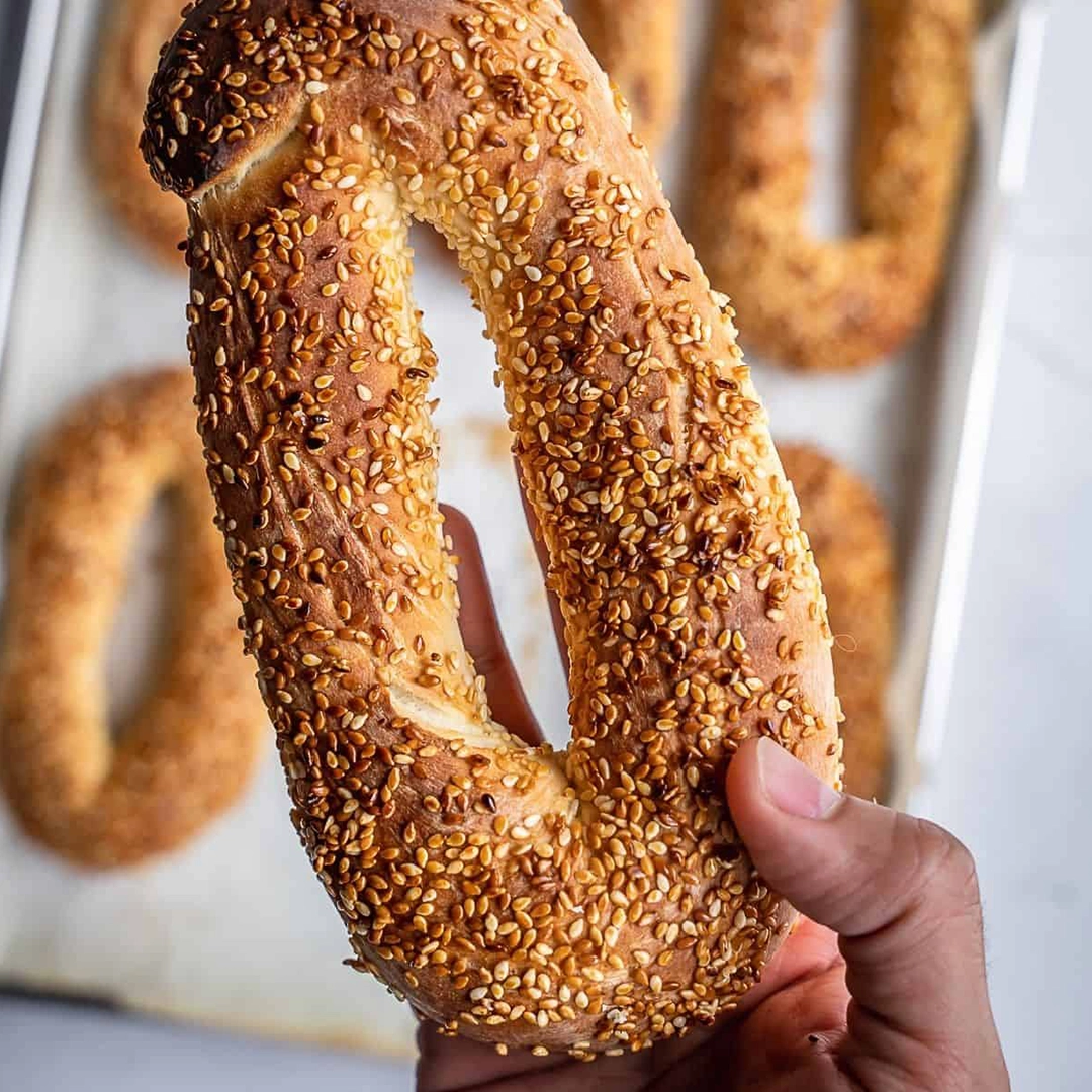 Hand holding a sesame-covered oval bread with more similar breads on a tray in the background.