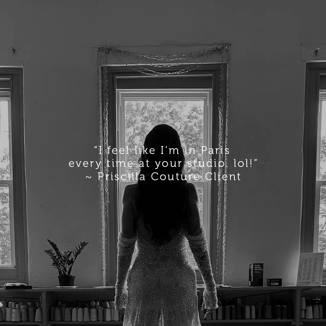Woman in a beaded dress standing with her back to the camera, looking out a window in a bright studio with spools of thread on shelves.