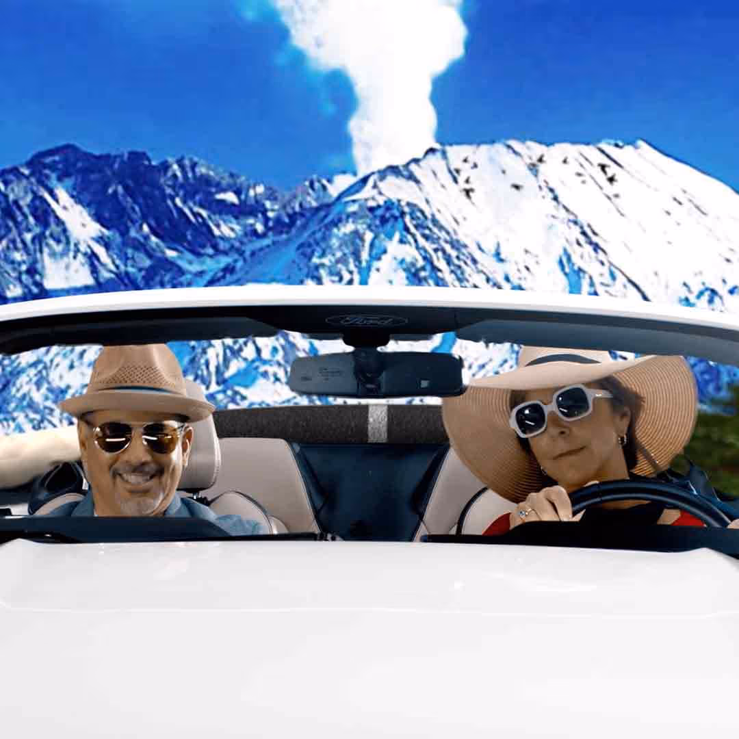 Man and woman wearing sunglasses and hats driving in a white convertible car with snow-covered mountains in the background.
